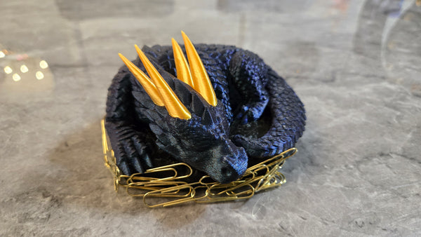 3D printed sleeping dragon in silk blue with gold horns curled in a black nest-shaped base used as a paper clip holder