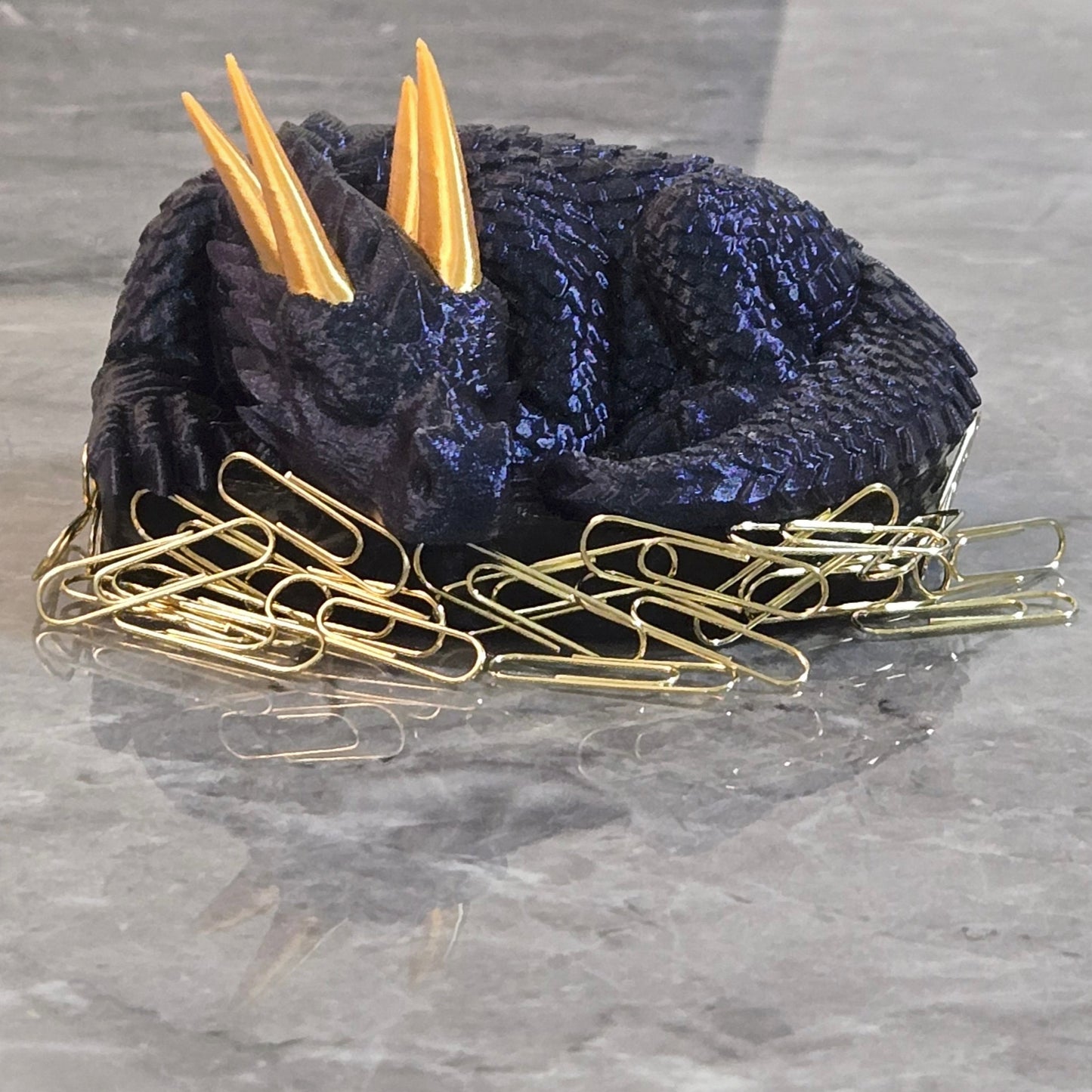 3D printed sleeping dragon in silk blue with gold horns curled in a black nest-shaped base used as a paper clip holder
